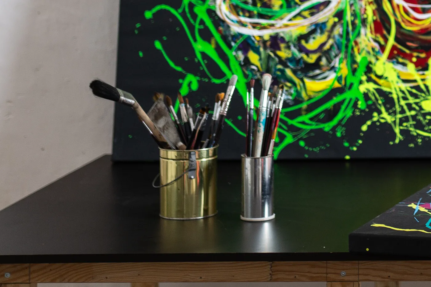 Two tins of used brushes on the black studio work table, with a painting of green and yellow looping lines behind them